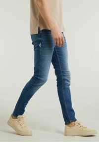 Jean en denim bleu slim avec des sections délavées, doté d'un design classique à cinq poches, associé à des baskets en toile beige.