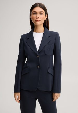 Femme portant un blazer bleu marine à deux boutons, une poche poitrine avec un détail brodé et un pantalon bleu marine assorti sur un top blanc.