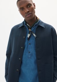 Jachetă bleumarin cu nasturi, cu guler, purtată peste o cămașă din denim, având un fular cu model legat la gât.