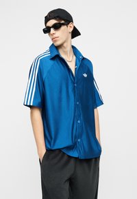 Młody mężczyzna w niebieskiej koszulce Adidas z białymi paskami, czarnych spodniach, czarnej czapce założonej do tyłu i czarnych okularach przeciwsłonecznych, z rękami w kieszeniach.