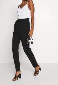 Svarta högmidjade byxor med knytbälte, tillsammans med en vit blus med volanger och svarta sandaler med remmar. Liten väska med ko-prickar.