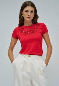 Camiseta roja de manga corta con diseño bordado floral en el escote. Combinada con pantalones blancos con pliegues, que presentan un detalle de botón.