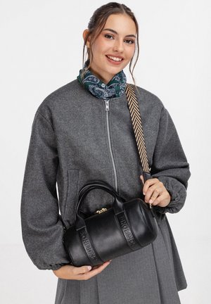 Lachende vrouw in een grijze jas met ritssluiting en een groene sjaal met patroon draagt een zwarte cilindervormige tas met een patroonband over haar schouder.