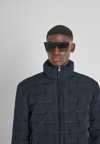 Giacca a vento nera con colletto alto, tessuto testurizzato con linee orizzontali. Indossata con occhiali da sole neri oversize.