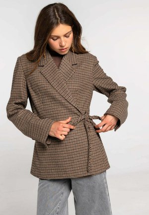 Abrigo de cuadros marrón con mangas largas, cuello de muesca y detalle de lazo en la cintura. Llevado sobre una blusa oscura y combinado con pantalones de pierna ancha gris claro.