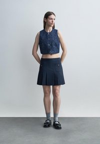 Γυναίκα που στέκεται με τα χέρια πίσω από την πλάτη, φορώντας ένα μπλε αμάνικο κοντό τοπ με κουμπιά, πλισέ σκούρα φούστα, γκρι κάλτσες και μαύρα παπούτσια.