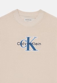 Bézs kötött pulóver, kerek nyakkal, bordás mandzsettával, és egy kék hímzett "CK" logóval, alatta "Calvin Klein" felirattal.