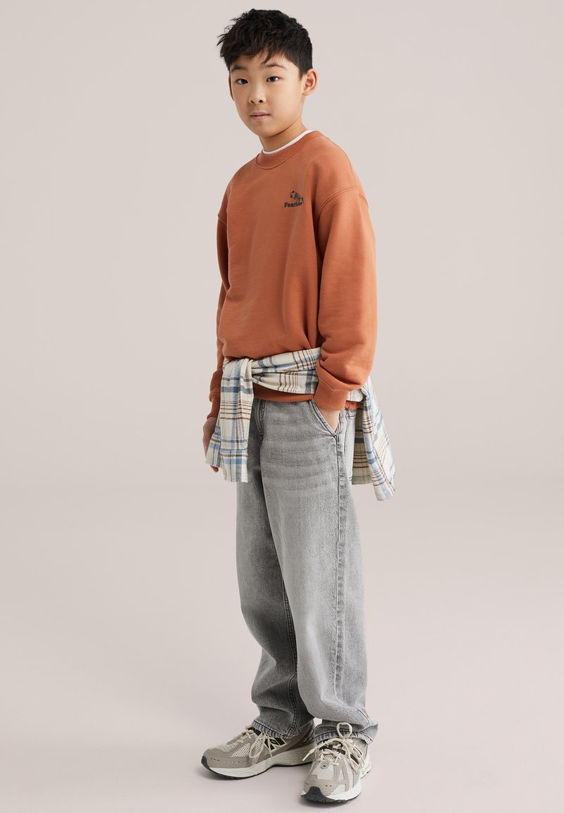 Sweat-shirt orange avec logo, chemise à carreaux nouée autour de la taille, jean gris ample et baskets grises avec accents. Cheveux courts noirs ; pose debout.