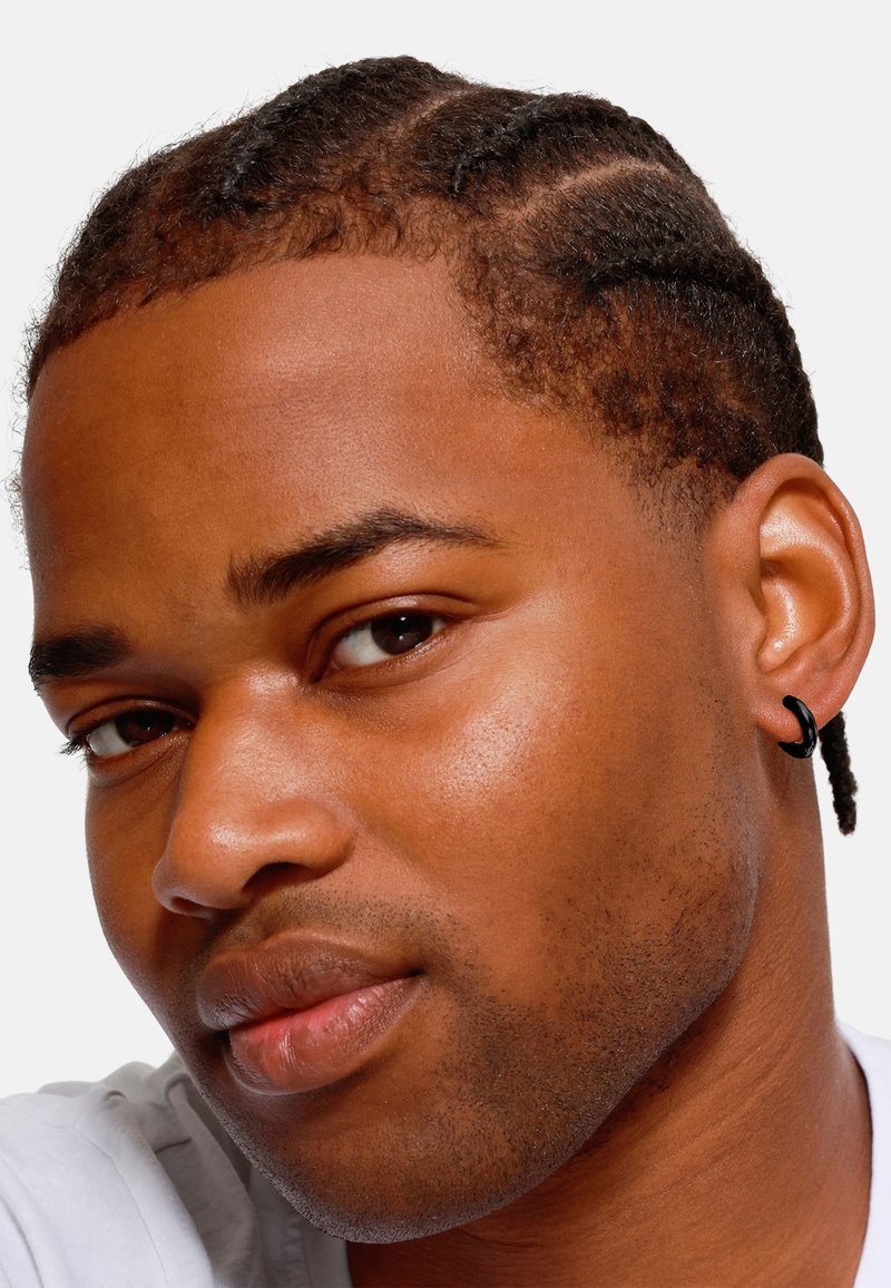 Nahaufnahme einer Person mit kurzen, geflochtenen Haaren, die einen schwarzen Creolenohrring trägt. Glatte Haut mit subtiler Textur und warmen Untertönen.