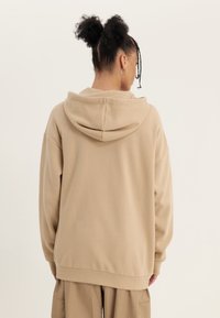 Felpa beige con cappuccio, dal fit rilassato, caratterizzata da una texture morbida, un ampio cappuccio e polsini a coste, indossata sopra pantaloni beige coordinati.