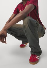 Červené sportovní boty s bílými pruhy, džíny a červený sportovní dres s černými akcenty. Tkanina je hladká a střih je volný.