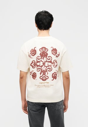 Mann trägt ein weißes T-Shirt mit kurzen Ärmeln, verziert mit braunem symmetrischem Blumendesign und Text auf der Rückseite, kombiniert mit dunkelgrauen Jeans.