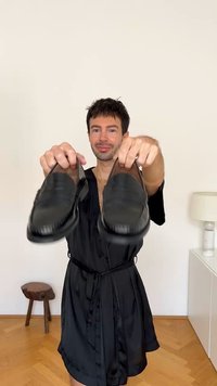 Schwarze Lederloafer mit glatter Textur, abgerundeten Zehen und dezenten Nähten, gehalten von einer Person in einem schwarzen Gewand.