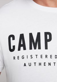 Camiseta blanca de algodón con texto negro en negrita que dice "CAMP REGISTRADO AUTÉNTICO," con cuello redondo y mangas cortas. Textura suave.