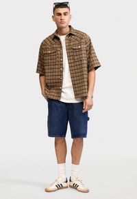 Mężczyzna z okularami przeciwsłonecznymi na głowie, w kraciastej koszuli z krótkim rękawem narzuconej na białą koszulkę, szorty dżinsowe, białe skarpetki i białe trampki z czarnymi paskami.