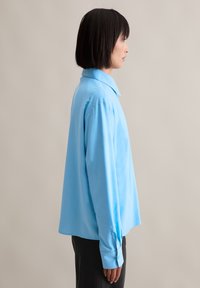 Light blue collared shirt with a loose fit, long sleeves, buttoned cuffs, and smooth texture, worn with dark pants.
