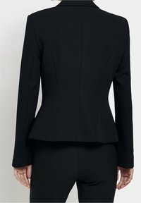 Bakifrån av en person som bär en åtsittande svart kavaj och matchande svarta byxor, stående mot en enfärgad ljus bakgrund.