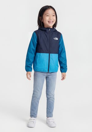 Usmievajúce sa dieťa v modro-námorníckej bunde na zips, svetlomodrých džínsoch a bielych teniskách, stojace pred jednoduchým pozadím.