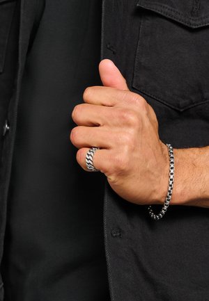 Hand mit silbernem Kettenring und passendem Kettenarmband, trägt eine schwarze Jacke mit sichtbarer Brusttasche und Knopf.