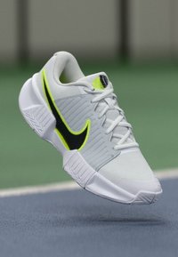 Λευκό αθλητικό παπούτσι με υφασμάτινη επιφάνεια, διαθέτοντας ένα μαύρο και νεον κίτρινο λογότυπο swoosh, στρογγυλή μύτη και καουτσούκ σόλα με μοτίβα πρόσφυσης.