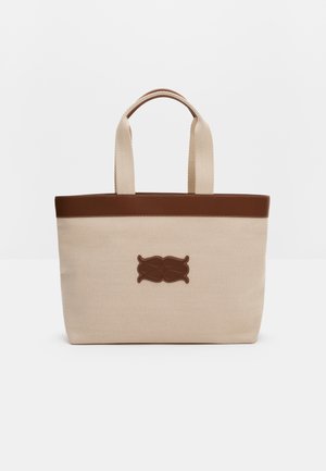 Sac fourre-tout en toile beige avec garniture en cuir marron, deux poignées et un emblème central en cuir marron sur fond blanc uni.