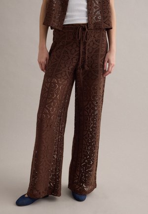 Donna che indossa pantaloni larghi marroni all'uncinetto con vita a coulisse, abbinati a un gilet coordinato e scarpe blu, in piedi su uno sfondo neutro.