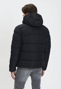 Geacă puffer neagră cu glugă, având un design matlasat și o textură izolată, prezentată din spate, purtată cu pantaloni din denim gri.