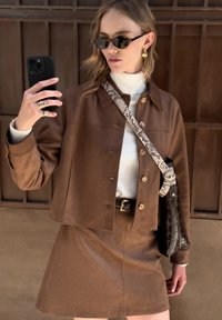 Chaqueta corta marrón con acabado texturizado, combinada con una falda a juego. Los accesorios incluyen gafas de sol y un bolso de mano con strap de patrón de serpiente.