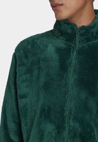 Chaqueta de forro polar verde con cuello alto, que presenta una tela suave y texturizada y una cremallera frontal. Adecuada para uso casual.