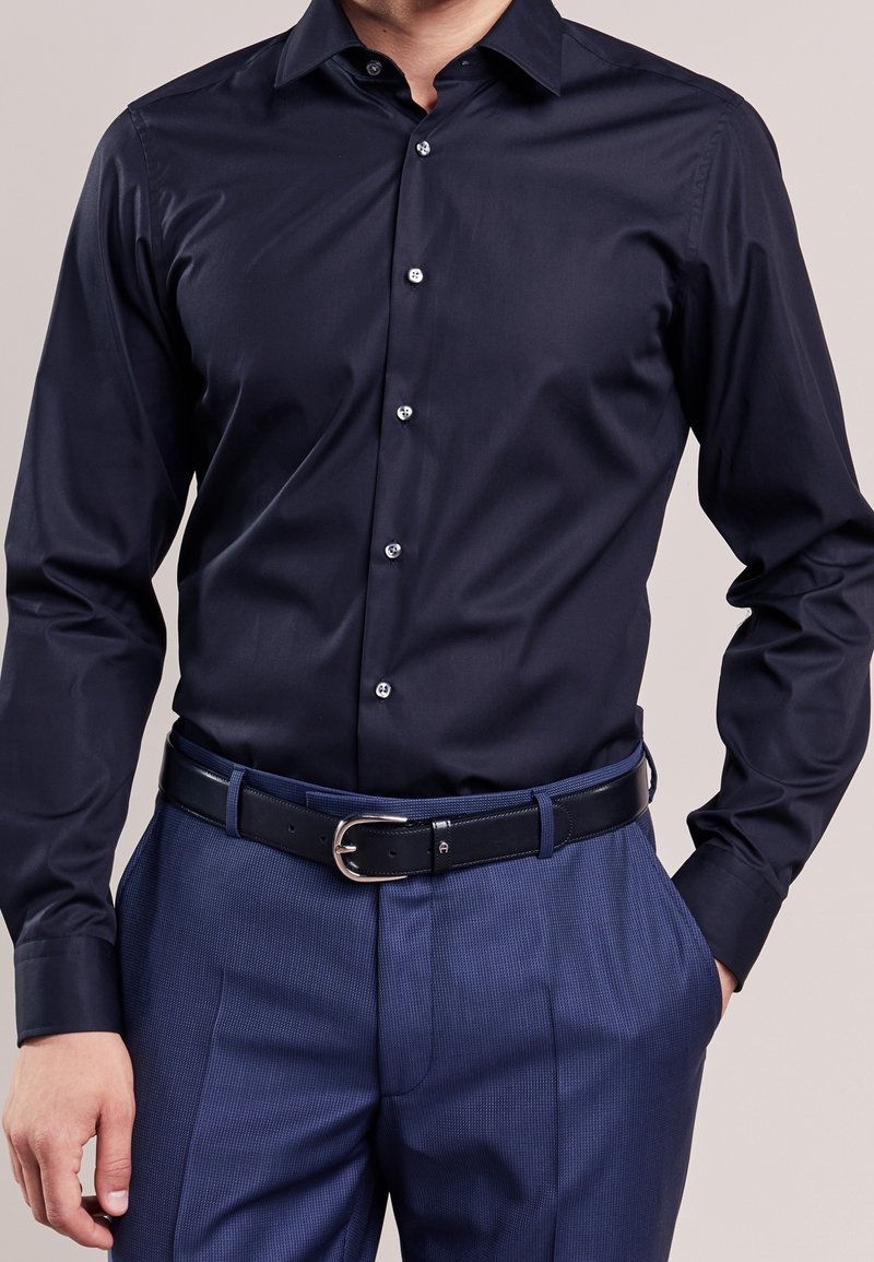 Homme portant une chemise ajustée bleu marine foncé rentrée dans un pantalon bleu texturé avec une ceinture en cuir noire.