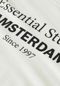 El texto en la tela blanca dice "Essential Stud AMSTERDAM Desde 1997" en fuentes negras con serifas y en negrita.
