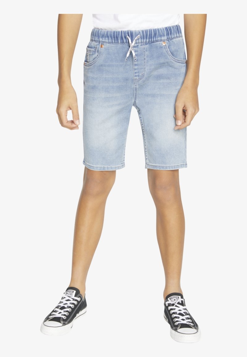 Garçon portant un short en jean bleu clair avec une taille élastique et un cordon blanc, associé à des baskets noires et blanches.