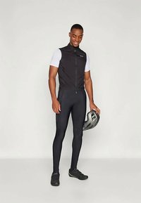 Black sleeveless cycling vest over a white short-sleeve jersey, paired with black leggings and shoes. Holding a gray helmet.