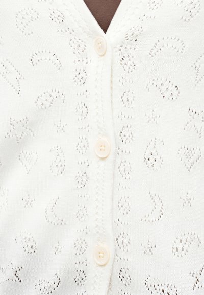 Λευκό πλεκτό ζακέτα με διάτρητα μοτίβα που περιλαμβάνουν καρδιές, αστέρια και σχήματα paisley, με κρεμ χρώματος κουμπιά στο κέντρο.