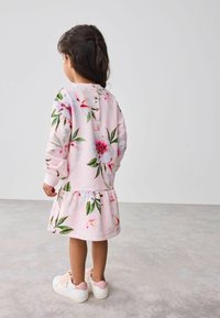 Rosa Blumenkleid mit langen Ärmeln, ausgestattet mit einem gerafften Saum, Knopfdetail am Rücken und grünen Blattmustern. Getragen mit weißen Sneakern.