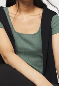 Grön ribbad kortärmad topp med fyrkantig halsringning, lagd under en svart tröja som hänger över axlarna. Slät textur, figurnära design.