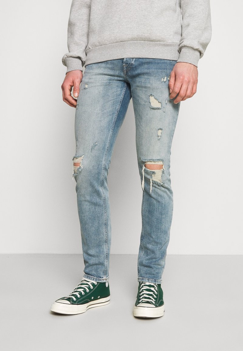 Ljusblå slitna jeans med rivna detaljer vid knäna, kombinerat med gröna högklackade sneakers. Materialet är denim med en urtvättad finish.