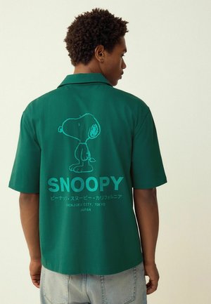 Grünes Kurzarmhemd aus Baumwolle. Auf der Rückseite befindet sich eine Linienzeichnung von Snoopy sowie der Schriftzug "SNOOPY" und "Shinjuku City, Tokyo."
