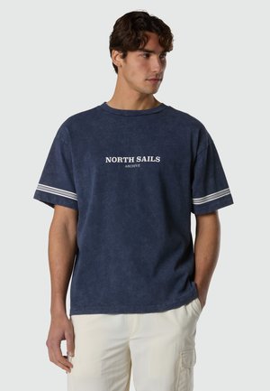 Hombre vistiendo una camiseta azul marino de manga corta con el texto "NORTH SAILS ARCHIVE" y detalles de rayas blancas en las mangas, combinada con pantalones de color claro.
