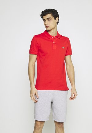 Polo rosso con colletto e maniche corte. Pantaloni atletici grigi. Logo Lacoste ricamato in verde sulla maglietta.