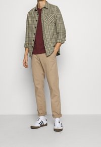 Camicia in flanella a quadri verde e marrone, maglietta bordeaux, pantaloni beige e sneaker bianchi con strisce nere. Outfit casual con vestibilità comoda.