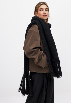 Zwarte franjesjaal gemaakt van textuurstof, hangend over een bruin oversized jasje. Zacht, warm uiterlijk met lange, ongelijke franjes.