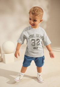 Gray short-sleeve T-shirt with a black graphic "ATHL. DEPT 32" paired with blue shorts. The child wears white sneakers and has curly hair.