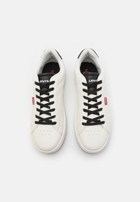 Sneakers in pelle bianca con lacci neri, caratterizzati da dettagli perforati e un logo Levi's sulla linguetta. Etichetta rossa a contrasto sul lato.
