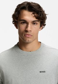 Szara bawełniana bluza z okrągłym dekoltem, z logo na lewej piersi, z prążkowanym wykończeniem na dekolcie i rękawach, noszona przez modelkę o kręconych włosach.