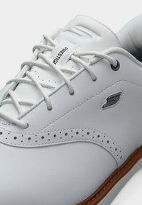 Baskets en cuir blanc avec lacets blancs, détails perforés, semelle marron et logo métallique "S" sur le panneau latéral.