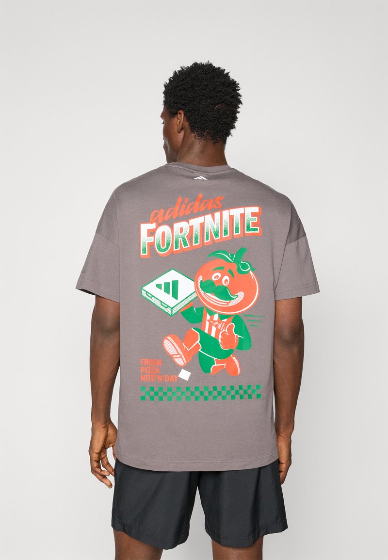 Szürke rövid ujjú póló, élénk grafika egy rajzfilmszerű paradicsom karakterrel, rajta a "adidas FORTNITE" felirattal és pixel dizájn elemekkel.