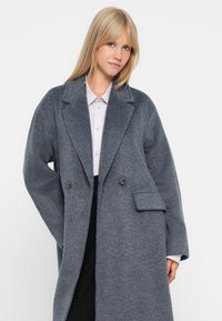 Grå uldblandingslang frakke med skræddersyet dobbeltbrystet design, brede søm, og en enkelt frontlomme. Glat tekstur, medium tykkelse.