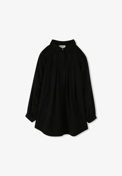 Blouse noire à manches longues avec un col structuré, présentant un empiècement avant orné de broderies en zigzag et des détails froncés au niveau des manches.
