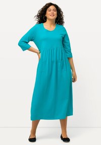 Błękitna sukienka z dżerseju z rękawami o długości ¾, okrągłym dekoltem i marszczoną talią. Posiada dwie przednie kieszenie i spódnicę do pełnej długości.